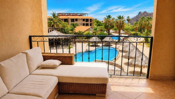 A balcony with a wicker sofa and a view of a pool area with palm trees and a building in the background.