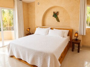 A large bed with white linens, two bedside tables with lamps, a decorative wall piece, and a balcony with curtains. - Ocean-view 1BR condo with pool, AC, kitchen, and washer/dryer. (Loreto)