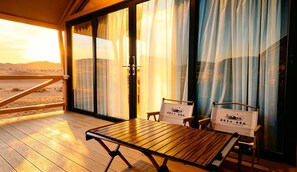 A room with a wooden floor, a table, two chairs, and a view of the desert through large windows with curtains.