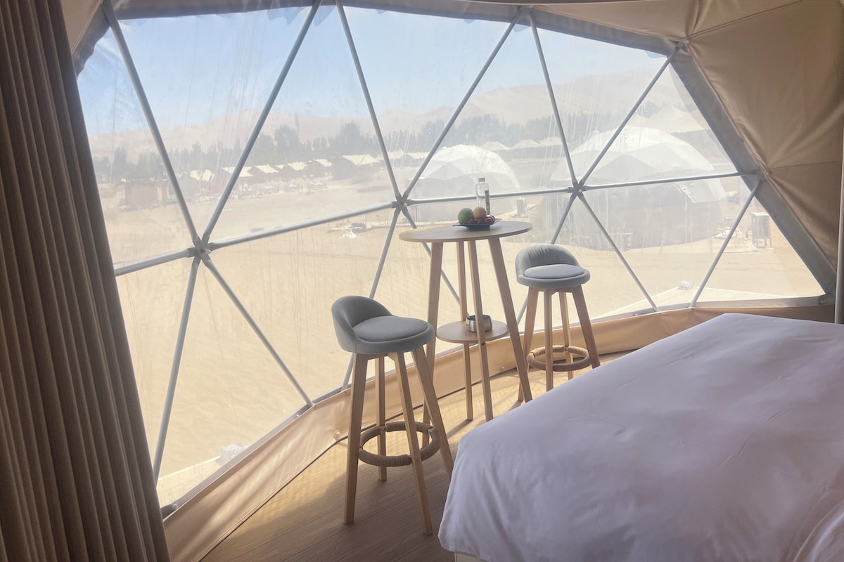 A tent with a bed, two stools, and a small table with a bottle and fruit.