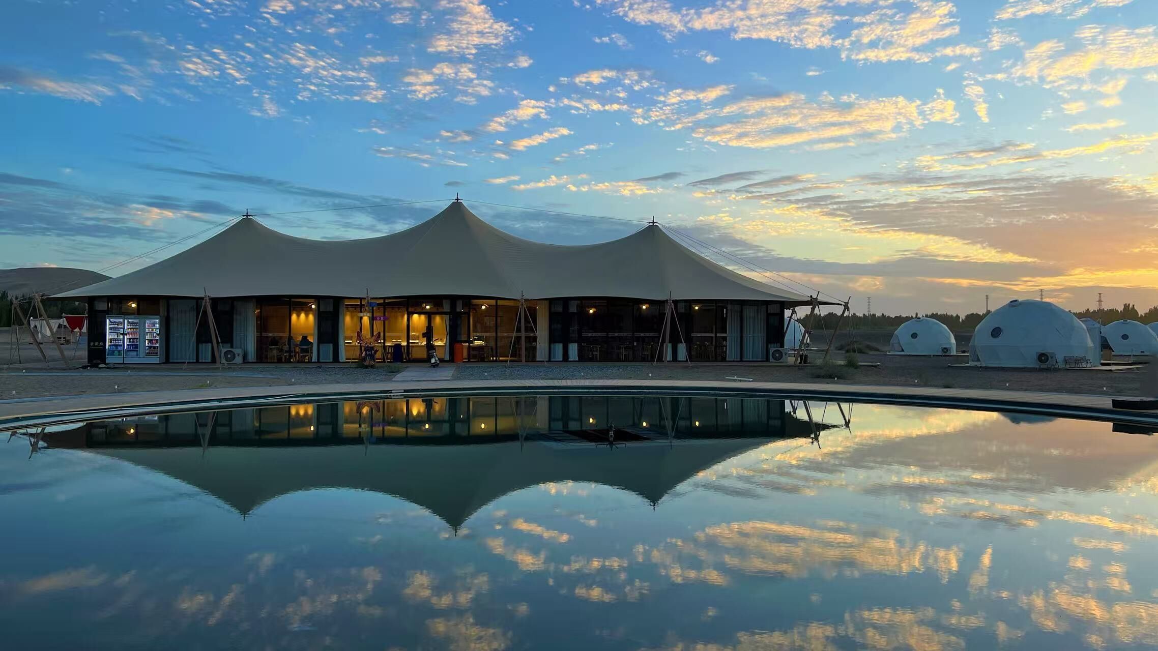 Un edificio moderno con piscina que refleja el cielo, rodeado de tiendas de campaña.