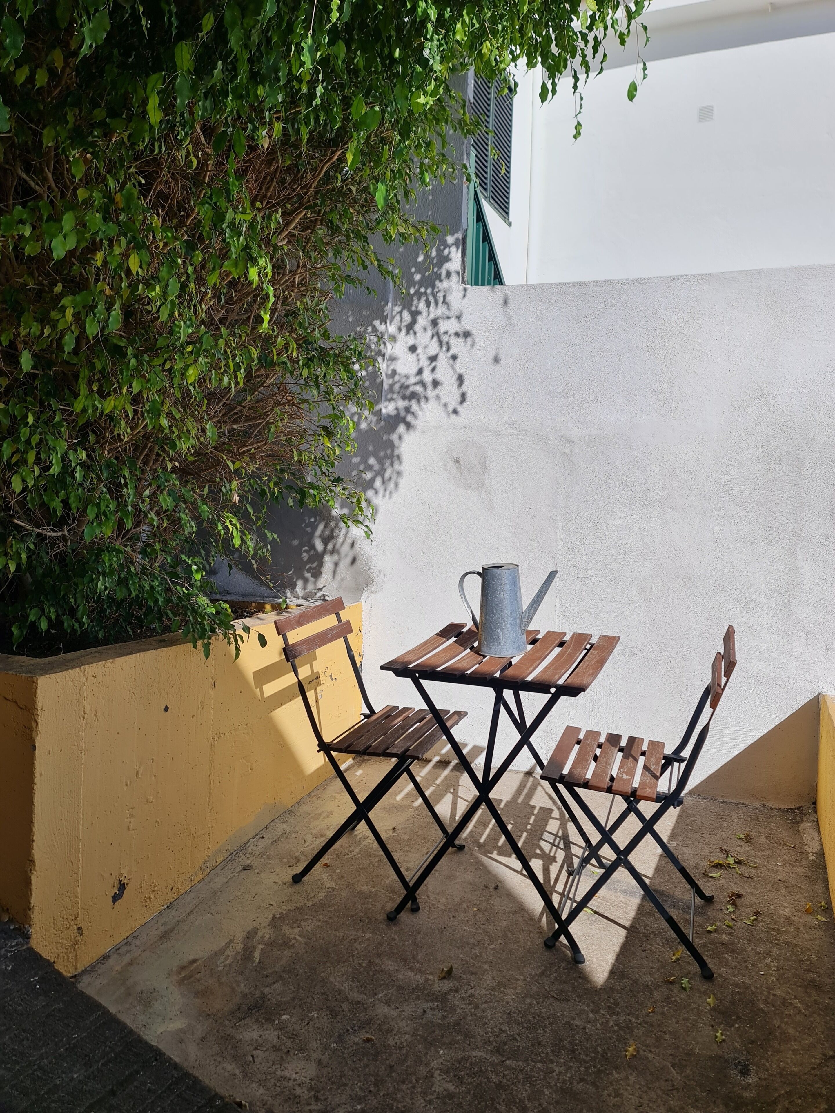 Een klein terras met een houten tafel en stoelen, een metalen gieter en een gele plantenbak met groene beplanting.
