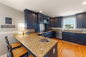 A modern kitchen with dark blue cabinets, granite countertops, and a large window offering a view. - Family Tree House | 2-Min Walk to Beach, Putting Green, Game Room & Large Fenced Yard (Holden Beach)