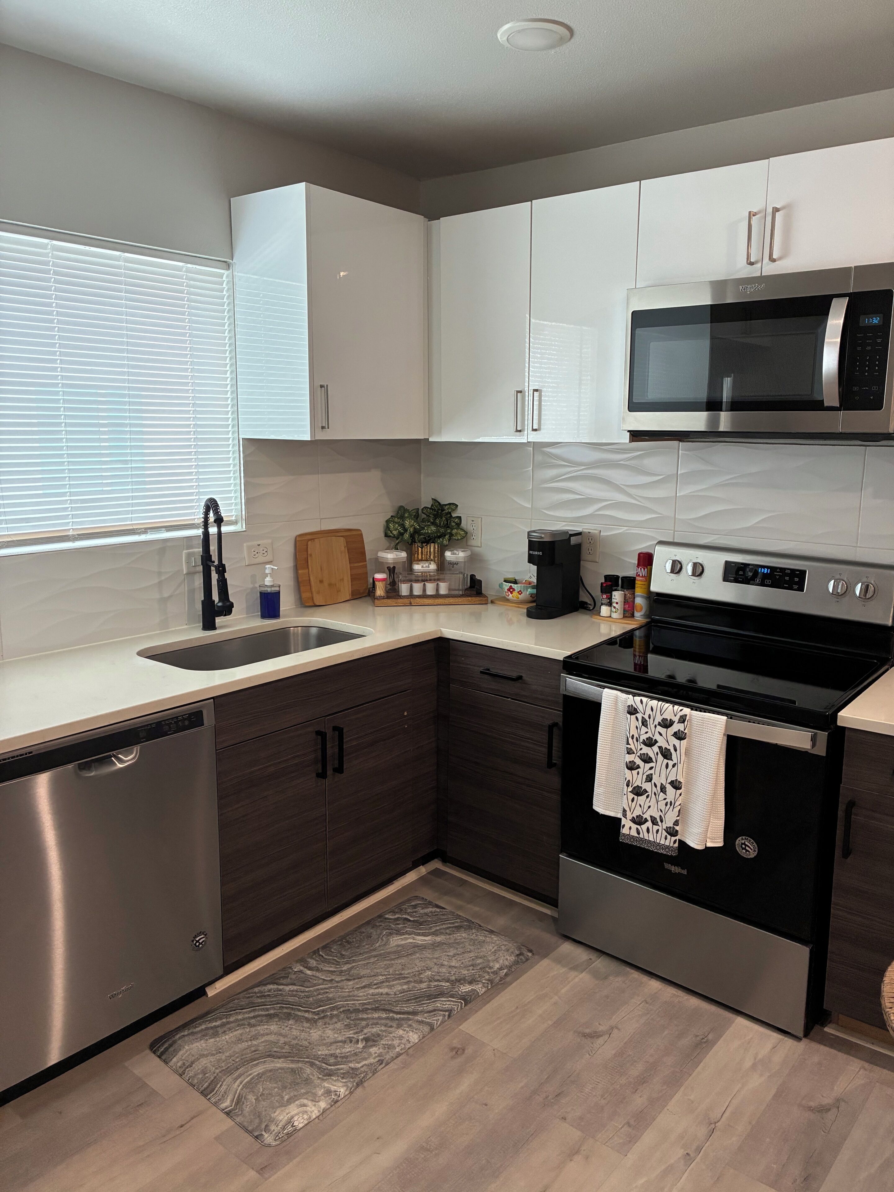 A modern kitchen with a dishwasher, stove, microwave, and a window with blinds.