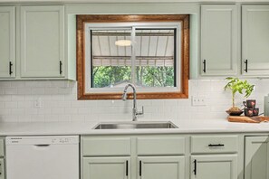 Una cocina con gabinetes blancos, un lavamanos de acero inoxidable, una ventana con marco de madera y una planta en maceta sobre la encimera.