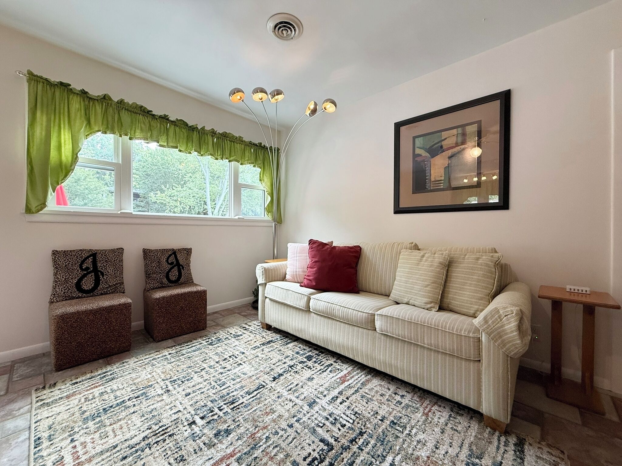 Una sala de estar con un sofá, dos sillones, un tapete estampado, una ventana con cortinas, una lámpara de techo e imagen enmarcada en la pared.