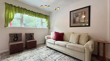 Una sala de estar con un sofá, dos sillones, un tapete estampado, una ventana con cortinas, una lámpara de techo e imagen enmarcada en la pared.