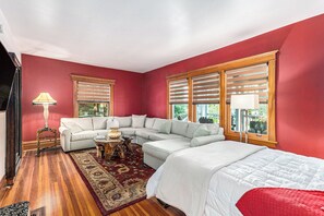 Quarto com sofá modular, cama, piso de madeira e grandes janelas com persianas.
