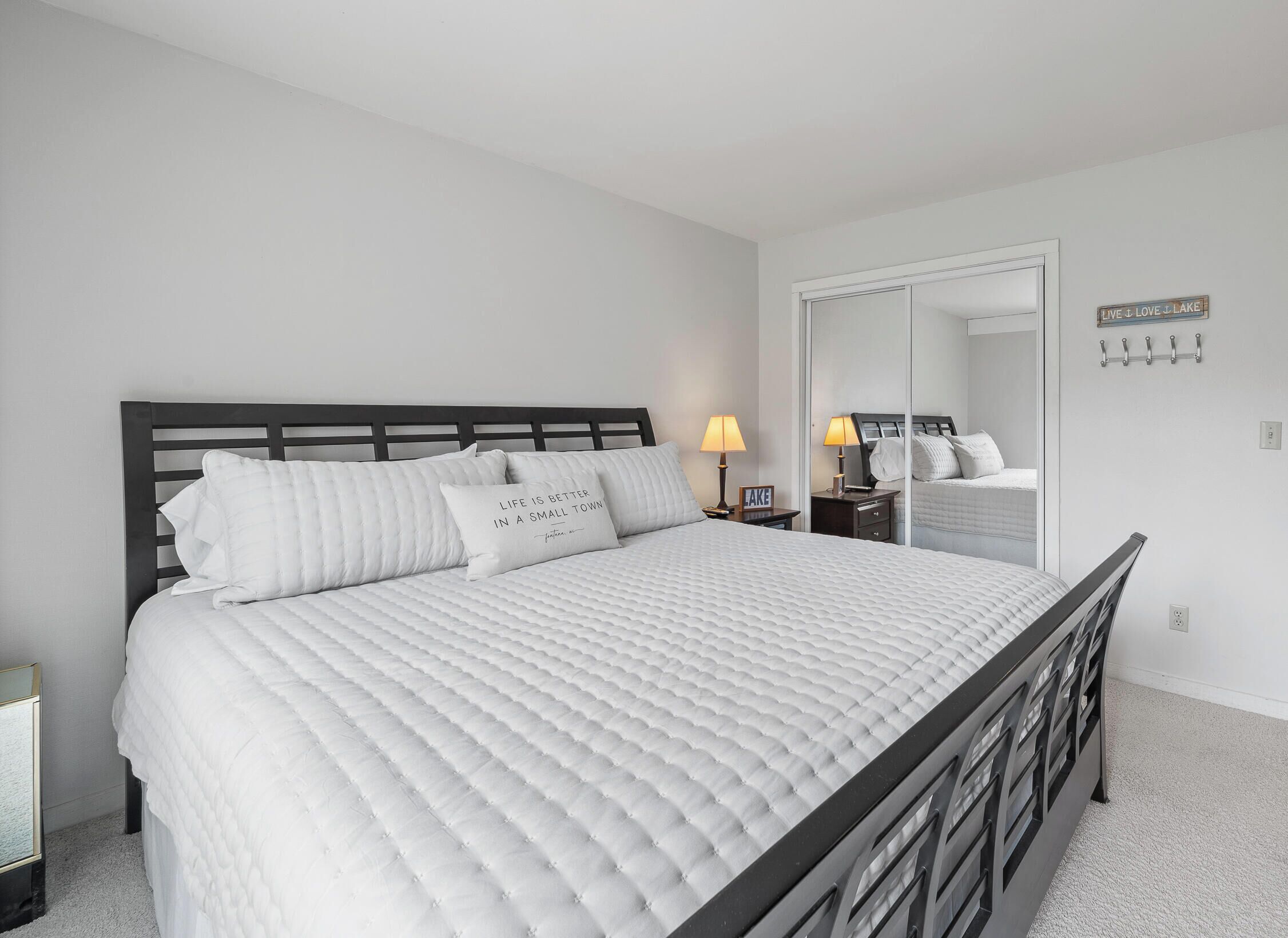 Cama bem arrumada com edredom branco acolchoado, cabeceira de madeira escura, duas luminárias de mesa e um espelho na parede.