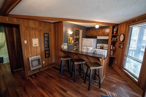A kitchen with wooden walls, a bar stool area, and a clock on the wall. - Charming Cabin by the Lifts — Walk or Shuttle in Minutes (Angel Fire)