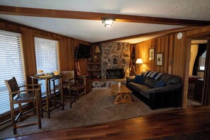 A cozy living room with wooden walls, a stone fireplace, a leather sofa, and a wooden coffee table. - Charming Cabin by the Lifts — Walk or Shuttle in Minutes (Angel Fire)
