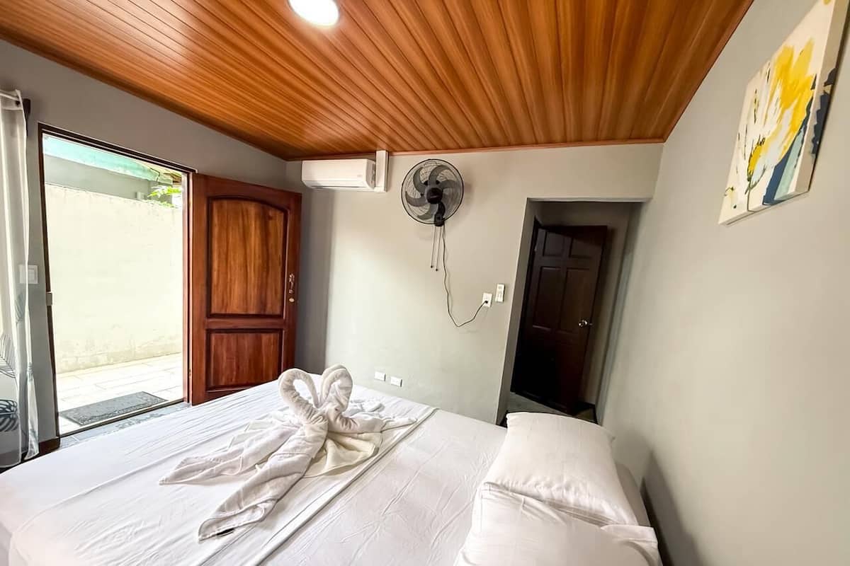 Ein Hotelzimmer mit Holzbalkendecke, einem Bett mit weißen Leinentüchern, einem Ventilator und einer Tür zu einem Balkon.