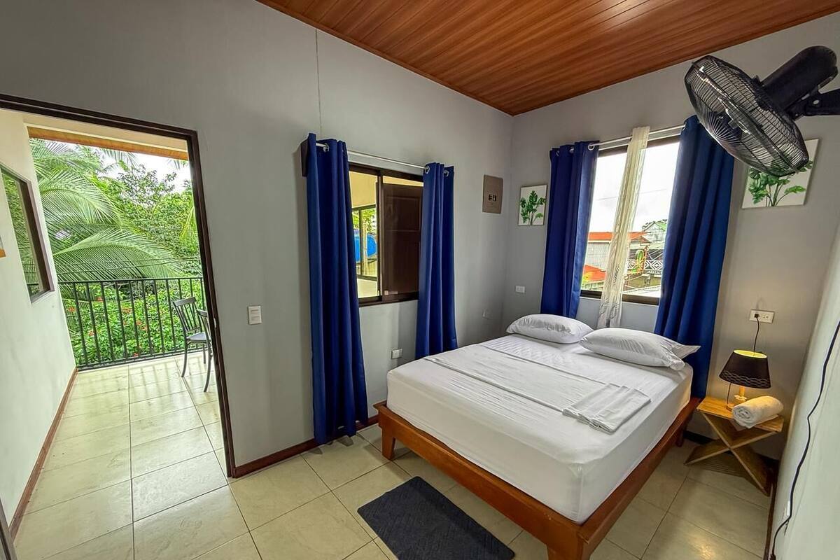 Ein Schlafzimmer mit Bett, Ventilator, Nachttisch und Blick auf einen Balkon.
