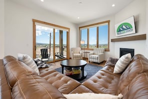 Ein geräumiges Wohnzimmer mit einer Ledersofa, gestreiften Sessel, einem Couchtisch und einem Kamin. Große Fenster bieten einen Blick ins Freie.