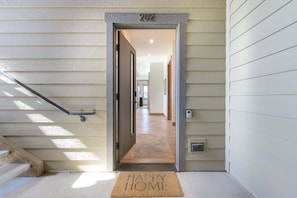 Ein Hauseingang mit der Hausnummer 202 über der Tür, einer Fußmatte mit der Aufschrift 'HAPPY HOME' und einer Treppe links.