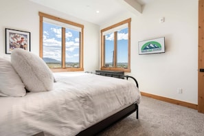 Ein Schlafzimmer mit einem Bett, zwei Fenstern mit Bergblick, einem gerahmten Bild und einem Logo an der Wand.
