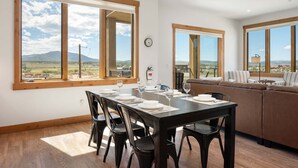 Un comedor con una mesa dispuesta para cuatro, ventanas amplias con vistas a montañas, un sofá y un reloj en la pared.