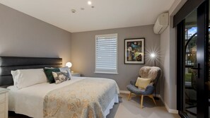 A bedroom with a bed, a chair, a nightstand, a window with blinds, a picture on the wall, and an air conditioning unit. - Apres Escape - 2 Bed (Queenstown)