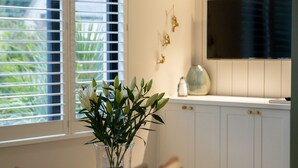 Ein Zimmer mit Fenster, einer Pflanze in einer klaren Vase, einem weißen Schrank mit goldenen Griffen und einem wandmontierten Fernseher.