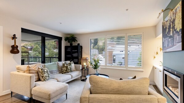 A living room with a sofa, armchairs, a fireplace, a large window with blinds, and a guitar on the wall. - Apres Escape - 3 Bed (Queenstown)
