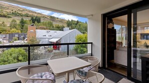 A balcony with a table and chairs, a view of a hillside, and a building with a metal roof. - Apres Escape - 3 Bed (Queenstown)
