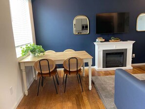 Een eethoek met een houten tafel en stoelen, een open haard, een aan de muur bevestigde televisie en een raam met luxaflex.