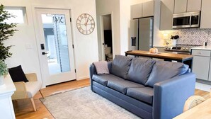 Una sala de estar moderna con un sofá gris, una mesa de centro de madera y una cocina con microondas y refrigerador.