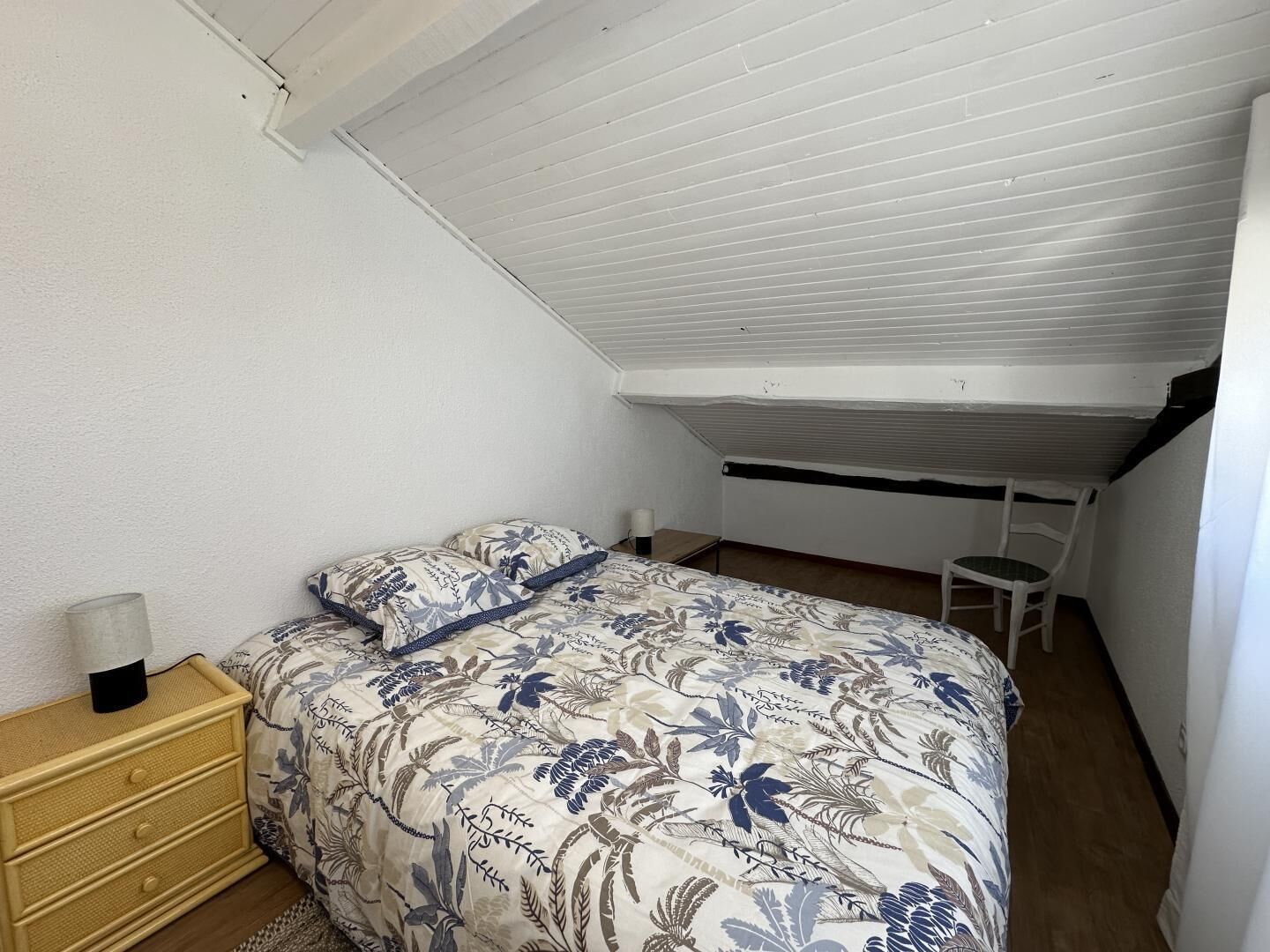 Un dormitorio con techo inclinado, una cama con ropa de cama floral, una mesita de noche, una silla y una pequeña mesa.
