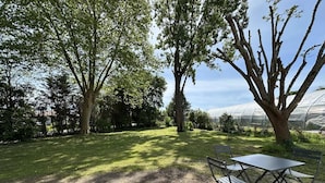 Eine Terrasse mit Tisch und Stühlen, umgeben von Bäumen und einem Gewächshaus im Hintergrund.