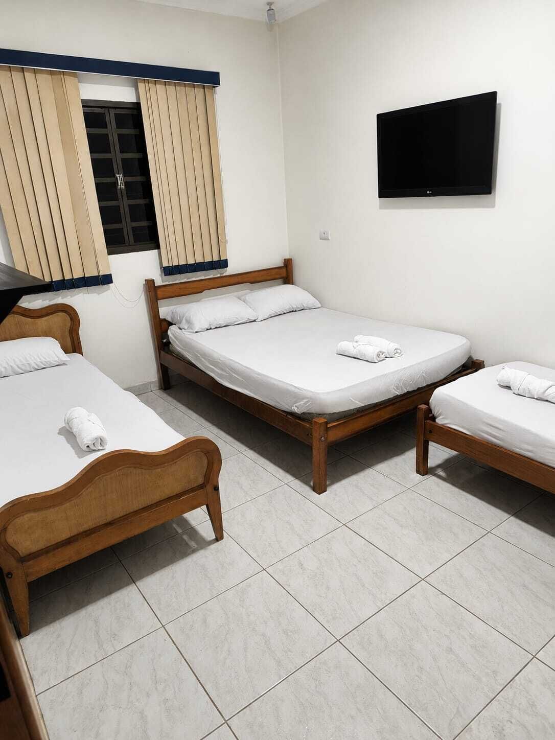 A room with three wooden beds, a flat-screen TV mounted on the wall, and a window with curtains.