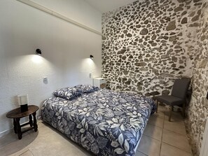 Een slaapkamer met een bed, een stoel, een klein tafeltje, een stenen muur en twee wandlampen.