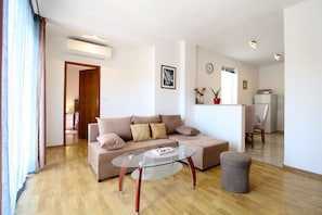Un salon comprenant un canapé, une table basse en verre, un plancher en bois, une porte, un climatiseur fixé au mur, une horloge, une petite plante et un réfrigérateur.