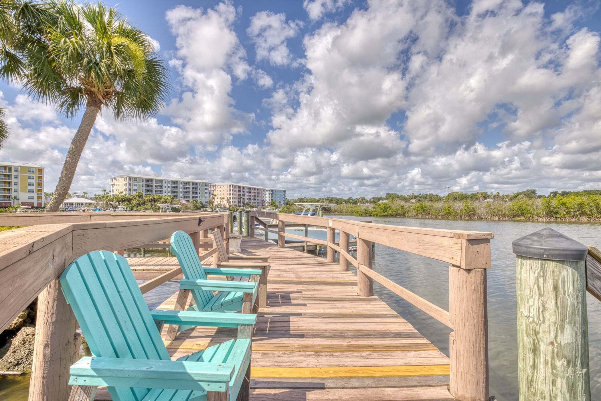 A wooden pier with chairs, a palm tree, and a view of buildings across the water.