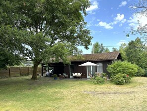 Ein rustikales Holzgebäude mit Ziegeldach, einer überdachten Terrasse mit Sitzgelegenheiten und einem großen Baum im Vordergrund.