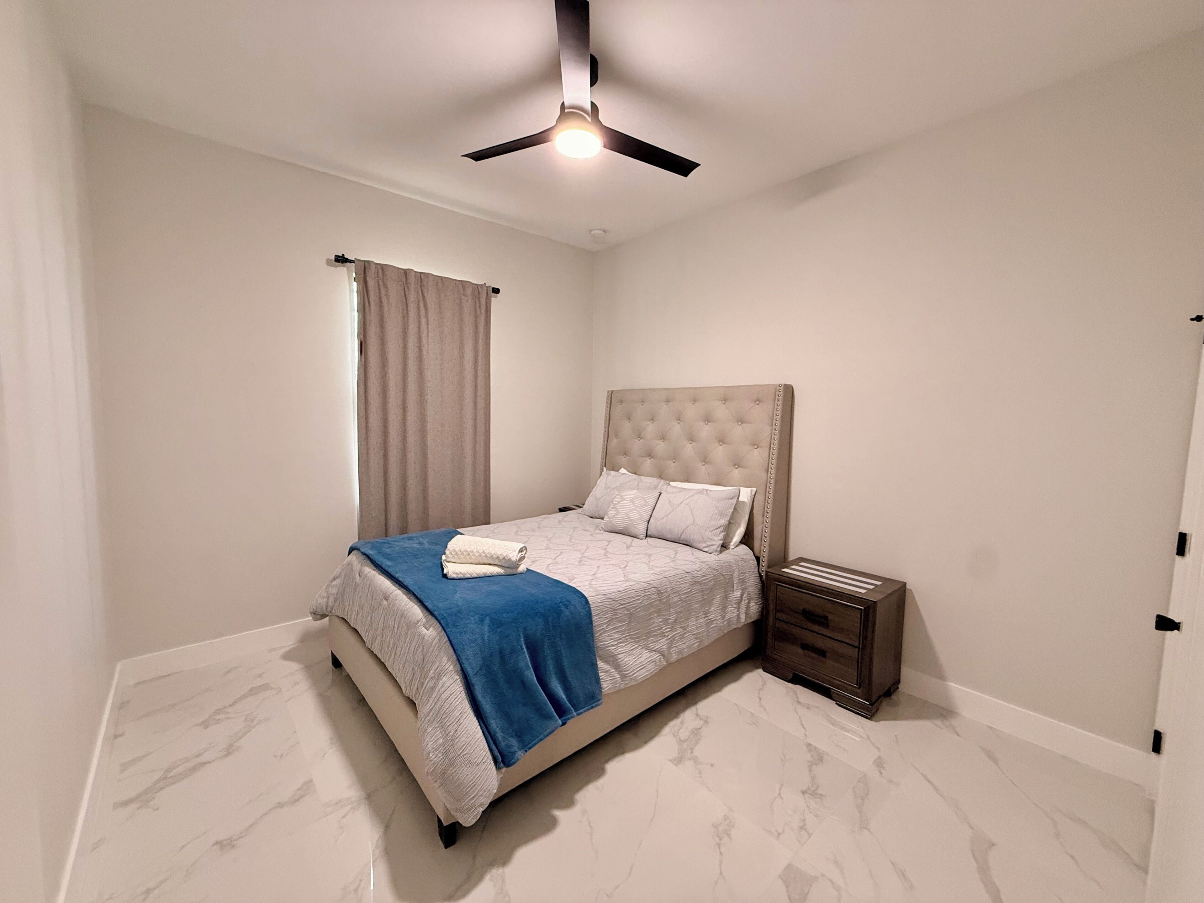 A bedroom with a bed, a nightstand, a ceiling fan, and a window with curtains.