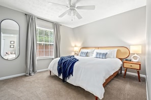Une chambre à coucher avec un lit, un ventilateur de plafond, une fenêtre avec des rideaux, une table de chevet avec une lampe et un miroir sur le mur.