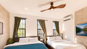Una habitación de hotel con dos camas, un ventilador de techo, un ventanal grande con cortinas, un televisor montado en la pared y un equipo de aire acondicionado.