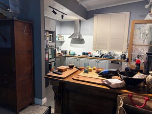 Une cuisine avec un plan de travail en bois, un luminaire suspendu, une fenêtre avec des stores et un réfrigérateur.