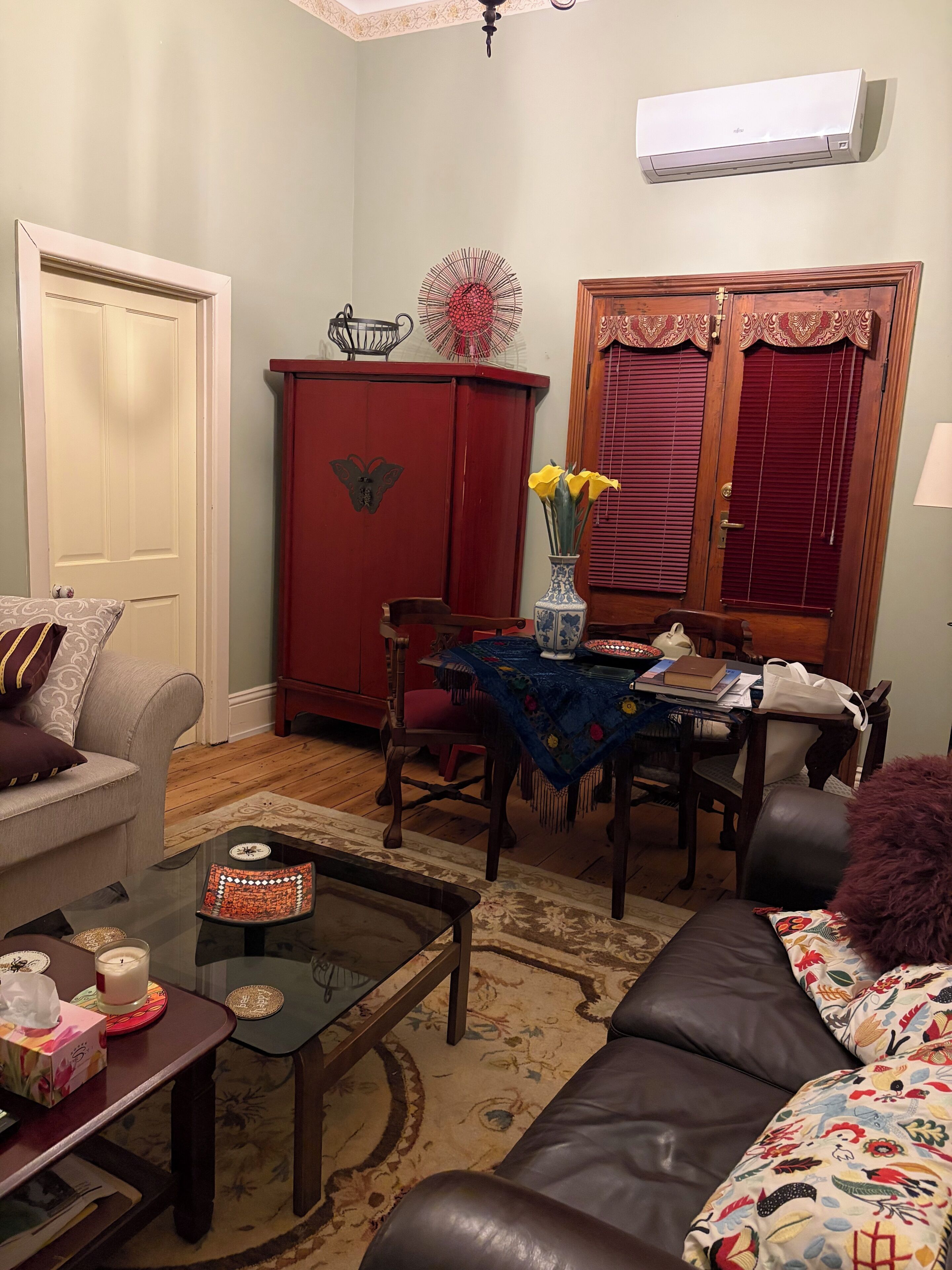 A living room with a sofa, a coffee table, a dining table with chairs, a red cabinet, and a window with shutters.