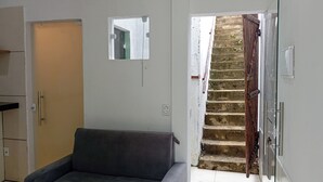 Una habitación con un sofá gris, una ventana pequeña, una puerta y una escalera que sube a un piso superior.