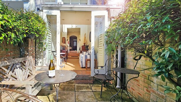 Eine Terrasse mit Tisch, Stühlen, einer Weinflasche und Blick in einen Raum mit Bett und Fenster.