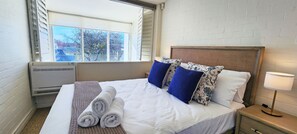 Een hotelkamer met een bed, een raam, een radiator en een nachtkastje met een lamp.