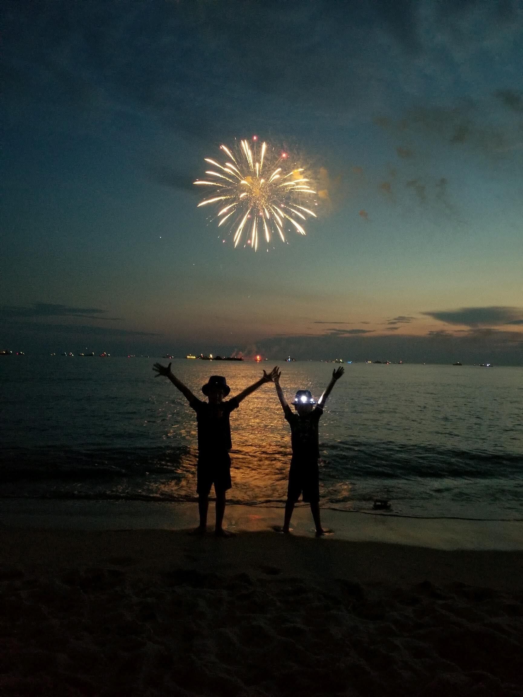 Deux silhouettes se dressent sur une plage, face à un spectacle de feux d’artifice qui illumine le ciel au-dessus de l’eau.