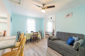 Um quarto com ventilador de teto, um sofá cinza com almofadas azuis, uma mesa de jantar com cadeiras e paredes azul claro.