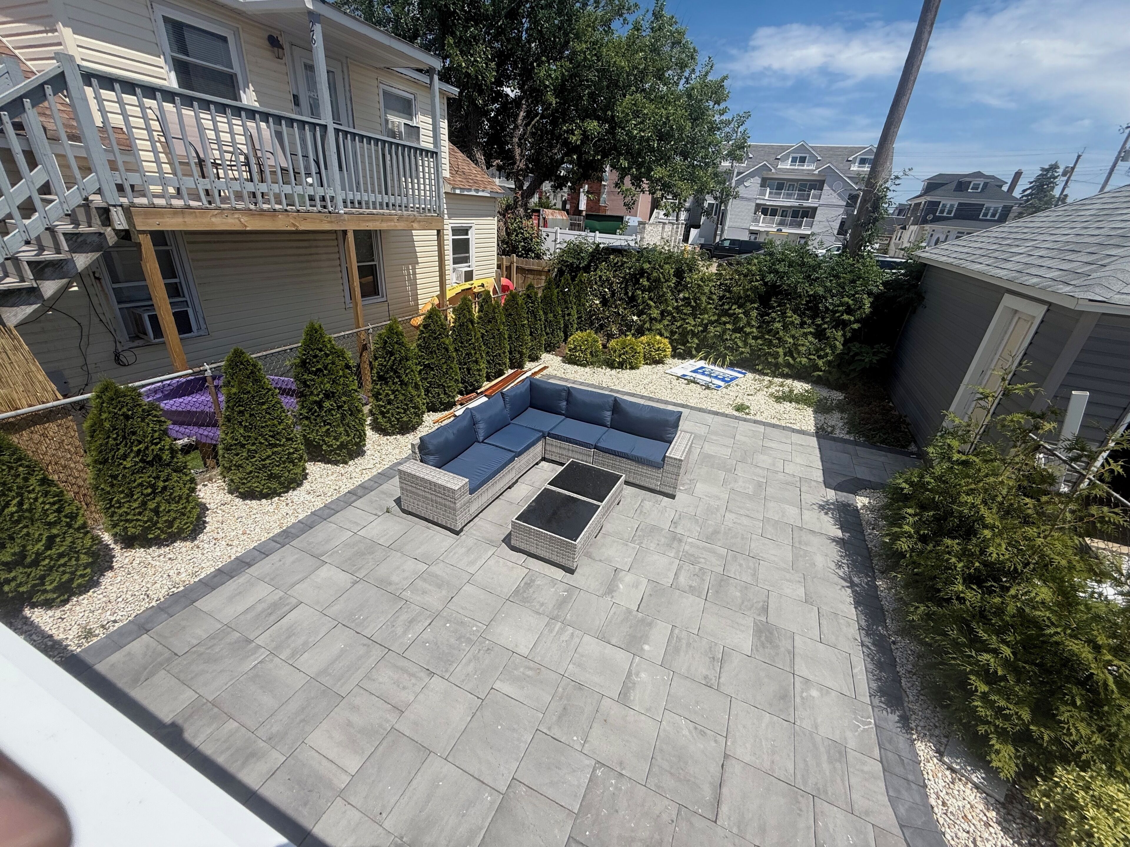 A patio with a sectional sofa and a rectangular table, surrounded by a gravel area and small trees.