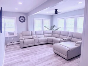 Ein modernes Wohnzimmer mit einer Eckcouch, einem Deckenventilator und großen Fenstern.