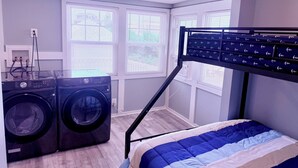 A laundry room with two front-loading washing machines, a bed with a blue and white striped comforter, and a window with blinds. - New! Tidal Escape - Beach & Boardwalk at Your Door (Seaside Park)