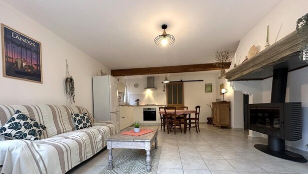 A living room with a sofa, a dining table with chairs, a kitchen in the background, and a fireplace.
