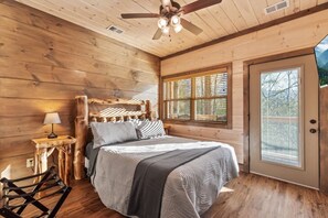 Une chambre à coucher dans une cabane en bois, comprenant un lit, une table de chevet, une lampe, un ventilateur de plafond et une fenêtre avec des stores.