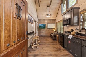 Ein rustikales Blockhaus-Interieur mit Holzwänden, einer Küche mit dunklen Schränken, einem Deckenventilator und einem Sofa im Wohnbereich.